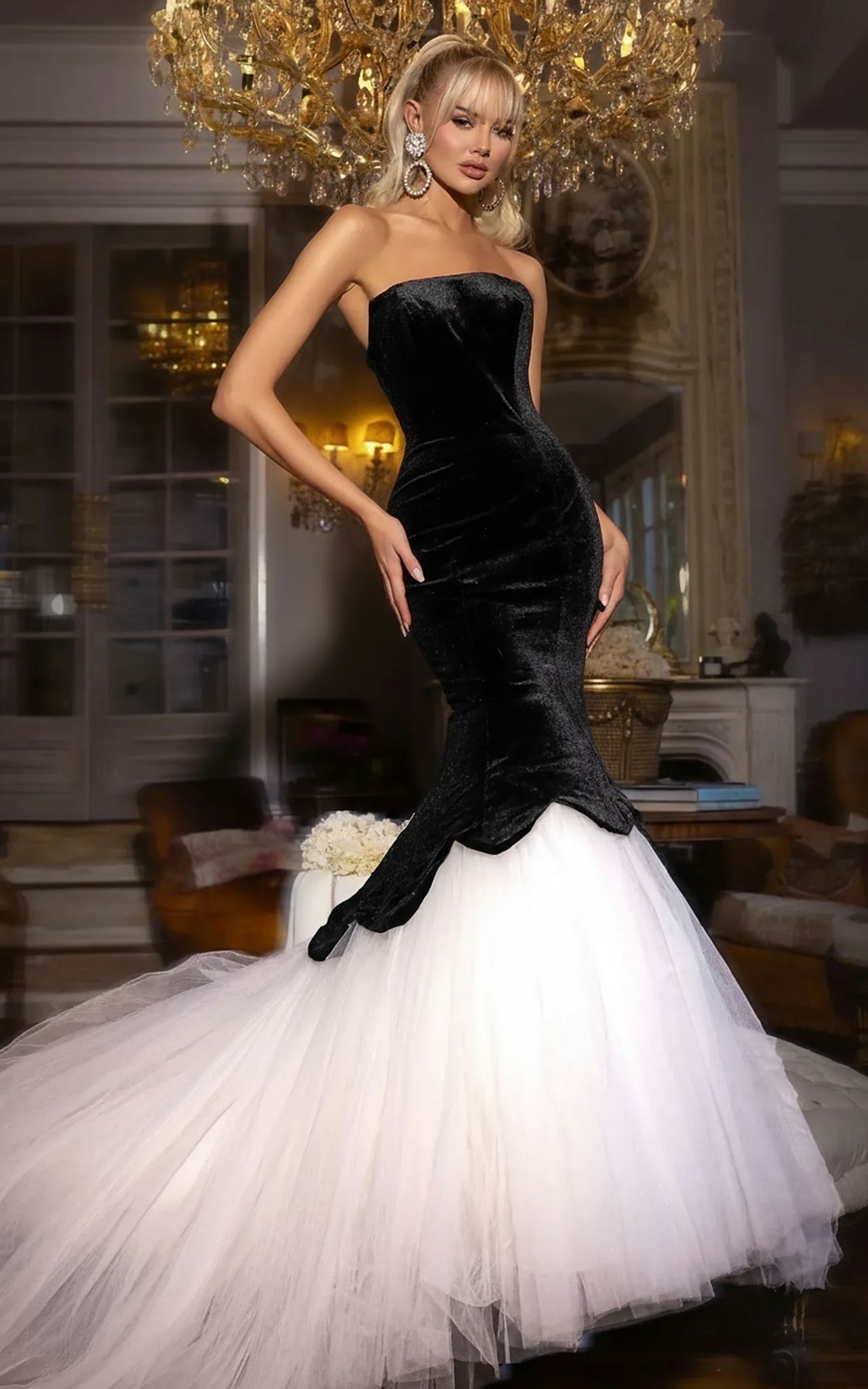 A woman stands in an elegant, chandelier-lit room wearing the Long Prom Dress: Portia and Scarlett PS25631E, a strapless mermaid gown in black velvet and white tulle.